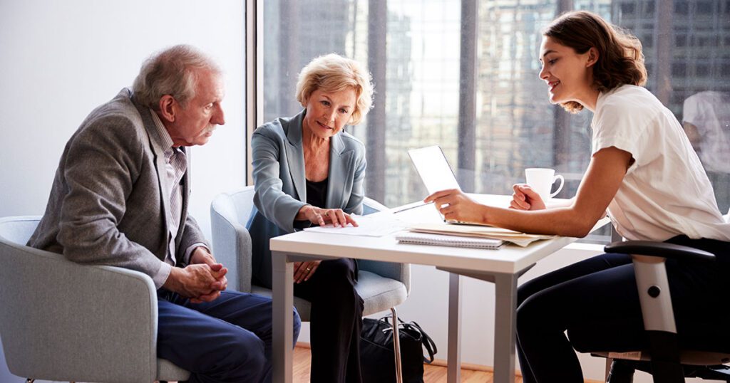 Older couple reviewing documents with a young female financial advisor at a table in a modern high-rise office, representing a conversation about investment diversification and portfolio planning