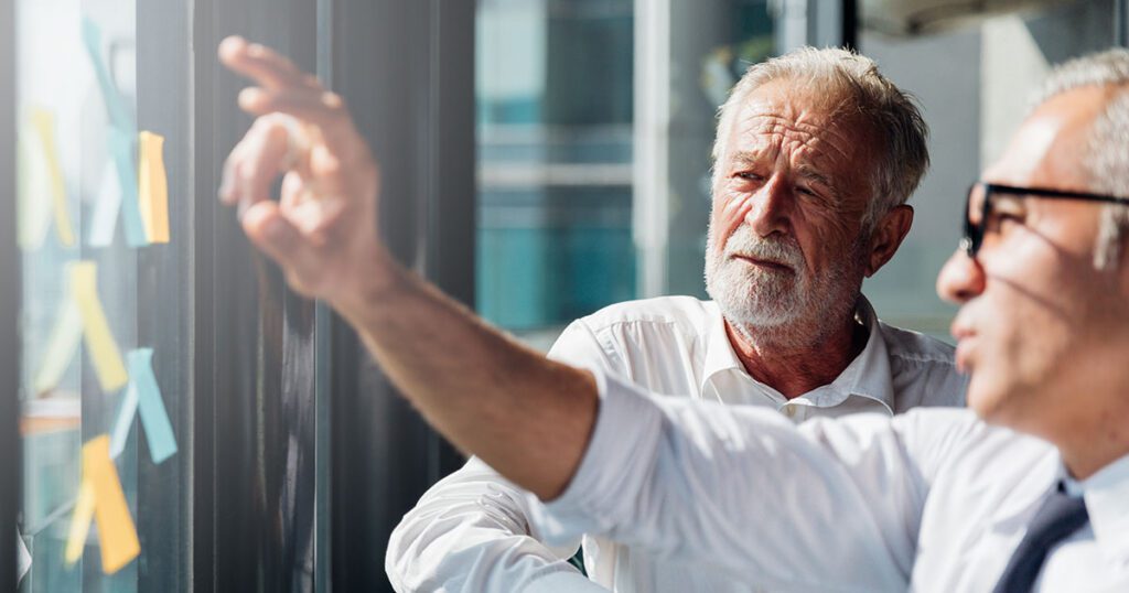 Two older professionals reviewing plans on a glass wall, representing how today’s millionaires take a more active, strategic approach to designing their retirement lifestyle