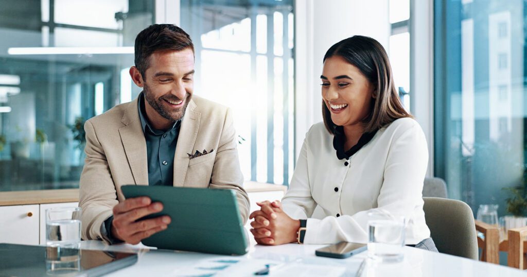 A male and female financial advisor smiling while reviewing wealth management strategies on a digital tablet in a modern corporate office.
