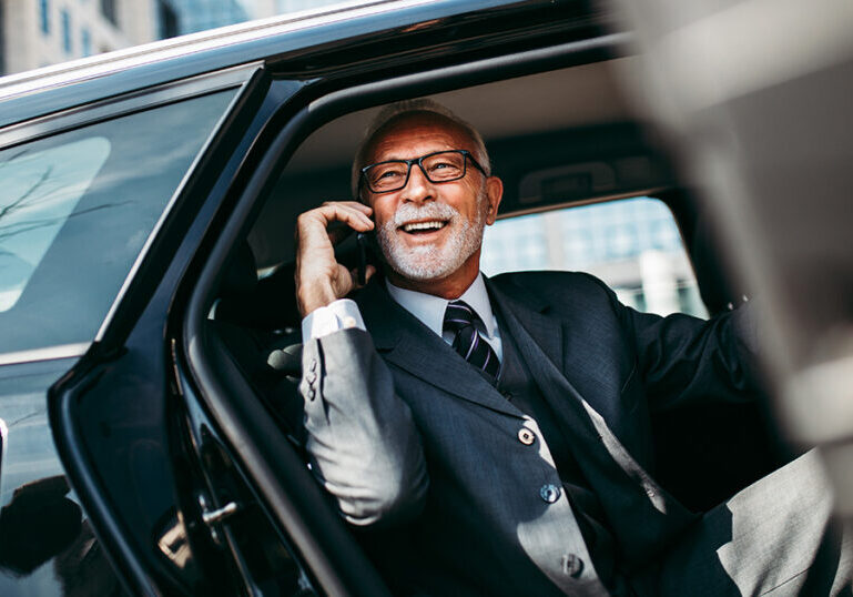 Good looking senior business man sitting on backseat in luxury car. He opens car doors and going or stepping out. Big business building in background. Transportation in corporate business concept.