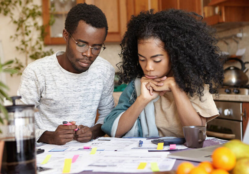 Young couple sitting at kitchen table with lots of papers, calculator and laptop