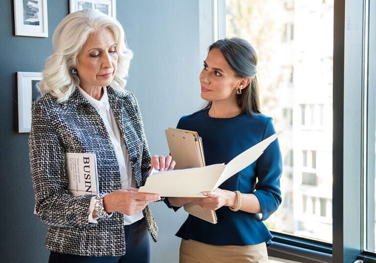 Senior female wealth advisor reviewing documents with a younger client in a high-rise office, representing tailored UHNW wealth management services for ultra-high-net-worth families
