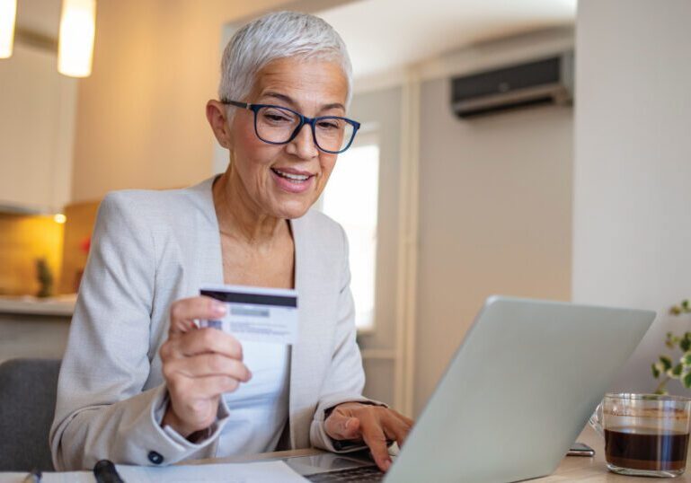 Woman reviewing financial information on a laptop and holding a credit card at her kitchen table