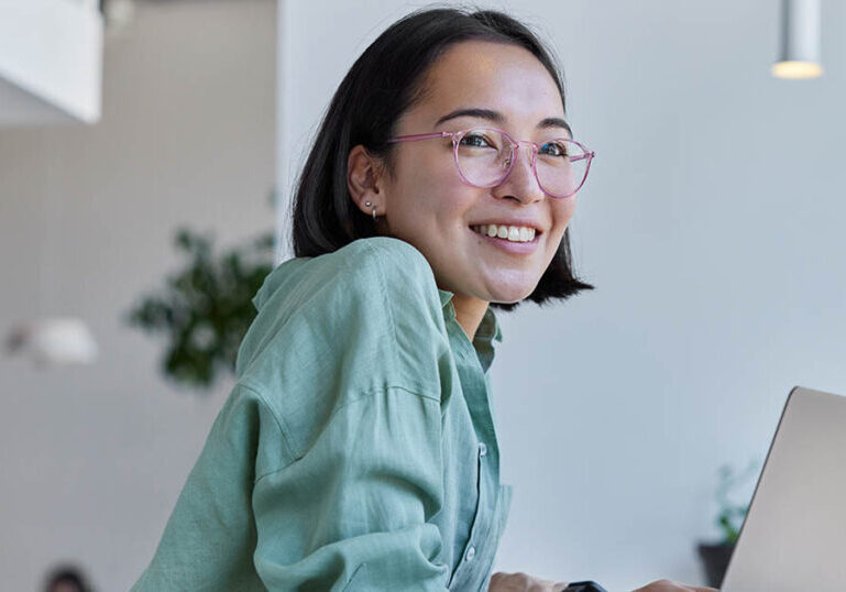 Pretty cheerful Asian woman in eyeglasses and casual clothes browses laptop computer connected to 4g internet updates software uses modern technologies poses in cafeteria looks gladfully away