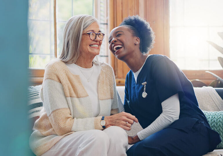 A smiling female healthcare professional in blue scrubs laughs warmly with an elderly woman wearing glasses and a cardigan while sitting together on a couch. The image represents the "Phase 3" long-term care stage discussed in the article, highlighting the importance of planning for personal home care needs in retirement.